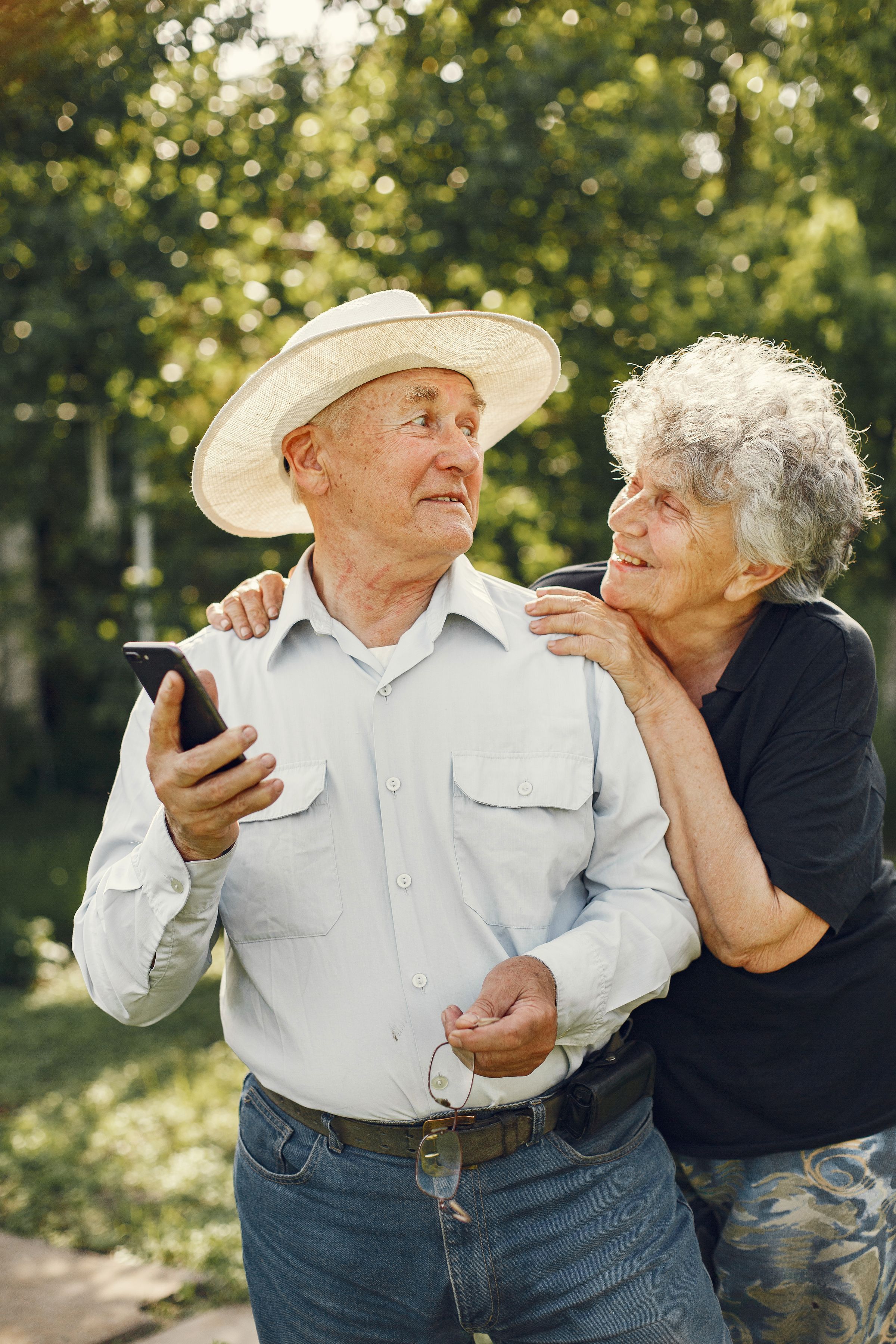 An older couple laughing together in a sunny garden, the man holding his phone.